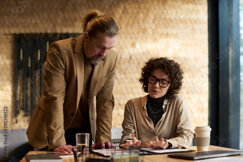Confident businesswoman explaining data in document to mature male colleague standing by her and bending over table during discussion