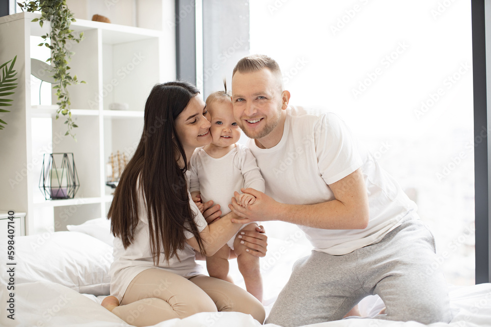 Cheerful caucasian family in casual attire sitting on white bed with charming baby girl and snuggling up to each other. Closeness during morning time of young parents and little daughter. Cheerful caucasian family in casual attire sitting on white bed with charming baby girl and snuggling up to each other. Closeness during morning time of young parents and little daughter.