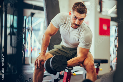A handsome muscular sportsman is sitting in a gym and doing exercises for biceps and triceps with a dumbbell.