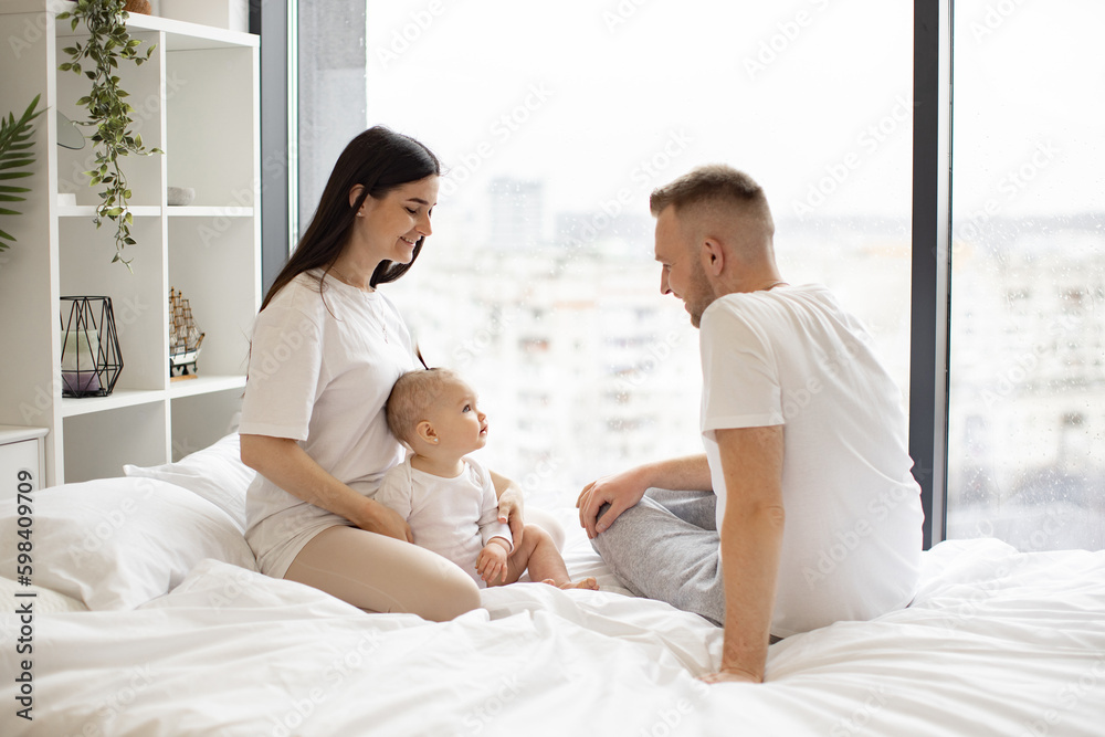 Obraz premium Cute little baby girl sitting together with loving parents on soft duvet in bedroom during weekend at home. Cheerful caucasian mom and dad taking care of pretty daughter while relaxing indoors.