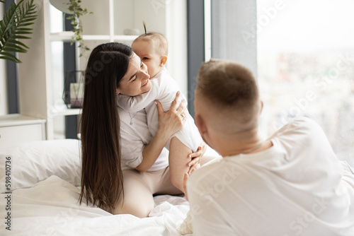Adorable baby infant hugging gently joyful brunette woman while happy blonde man lying on soft bed in studio apartment. Affectionate young family sharing love and closeness while living full life.