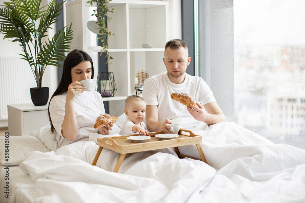 Beautiful woman and handsome man sitting under duvet and having drinks and snacks while cute baby taking dessert from bed tray. Young parents and daughter enjoying excellent food in bright interior.