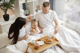 Top view of married caucasian couple helping baby girl eating roll while resting on bed with tray table in studio apartment. Loving young people taking care of cute daughter during morning time.