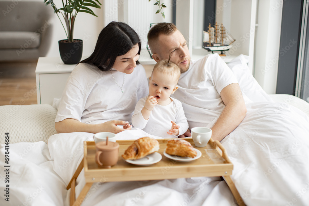 Sweet little baby drinking from sippy cup while joyful brunette woman snuggling with blonde male in cozy bedroom with tray on foreground. Caring family spending quality time together at home. Sweet little baby drinking from sippy cup while joyful brunette woman snuggling with blonde male in cozy bedroom with tray on foreground. Caring family spending quality time together at home.