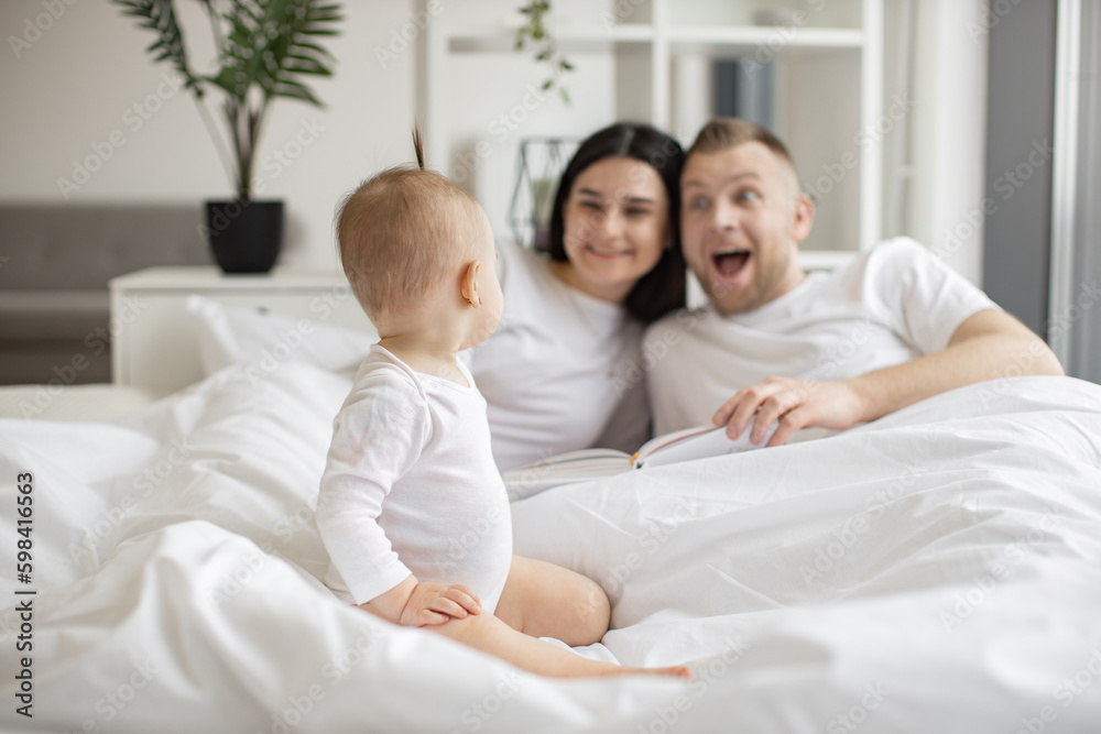 Focus on adorable little child sitting on white soft duvet and watching her parents playing roles of book characters on Sunday morning at home. Concept of family and togetherness. Focus on adorable little child sitting on white soft duvet and watching her parents playing roles of book characters on Sunday morning at home. Concept of family and togetherness.