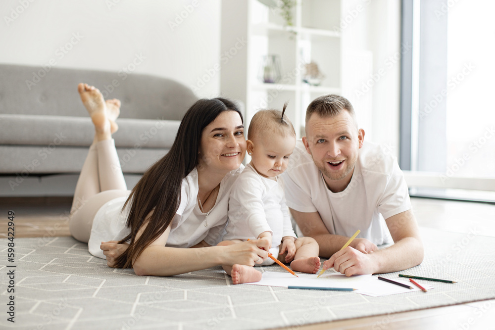 Smiling family of three in casual attire posing with drawing pencils while relaxing on wooden floor in studio apartment. Funny caucasian spouses developing baby's imagination using art and colors. Smiling family of three in casual attire posing with drawing pencils while relaxing on wooden floor in studio apartment. Funny caucasian spouses developing baby's imagination using art and colors.