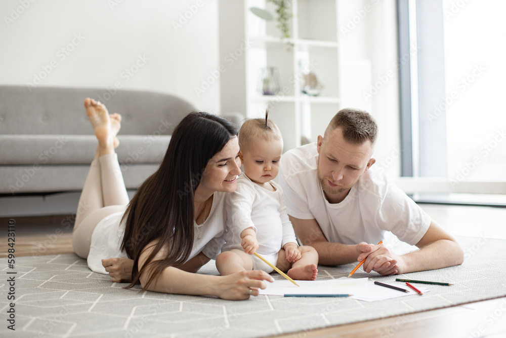 Adorable little baby girl in white cozy bodysuit holding red pencil while sitting on floor with mindful mother and father on both sides. Adult people and kid enjoying first scribbles on paper at home. Adorable little baby girl in white cozy bodysuit holding red pencil while sitting on floor with mindful mother and father on both sides. Adult people and kid enjoying first scribbles on paper at home.