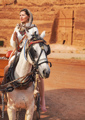 Young woman with white dress and head scarf, sitting on a horse, blurred Hegra or Mada’in Salih ruins in background - female tourist travel influencer like photo from Saudi Arabia