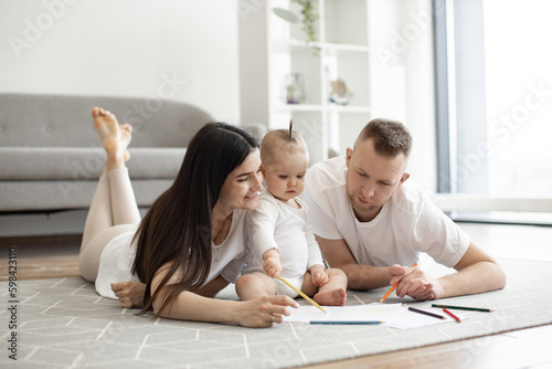 Adorable little baby girl in white cozy bodysuit holding red pencil while sitting on floor with mindful mother and father on both sides. Adult people and kid enjoying first scribbles on paper at home.