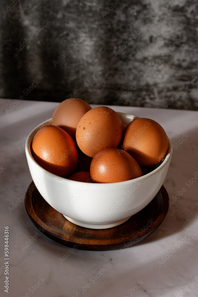Several brown chicken eggs in a white ceramic bowl, on white marble table against dark background Several brown chicken eggs in a white ceramic bowl, on white marble table against dark background