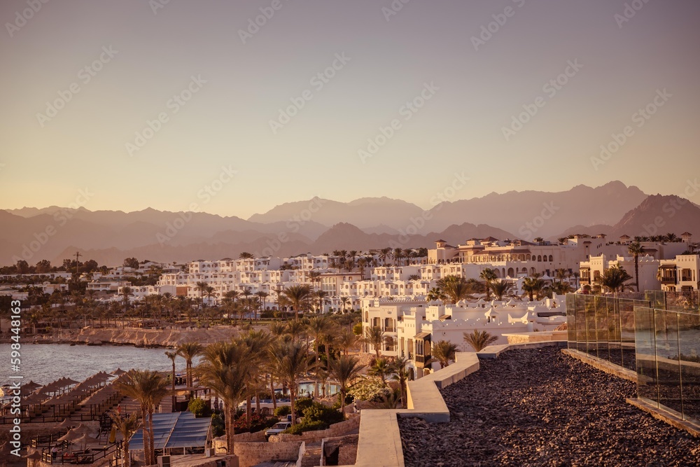 Imposing desert mountains provide a stunning backdrop to a cluster of white buildings, nestled amidst flourishing palm trees, capturing the essence of a tranquil desert oasis