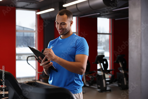 Happy trainer writing down workout plan in modern gym, space for text