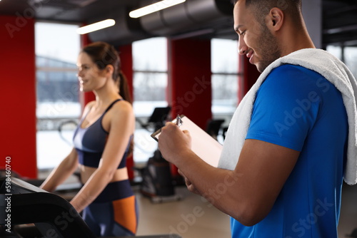 Happy trainer writing down workout plan while woman doing exercise in modern gym