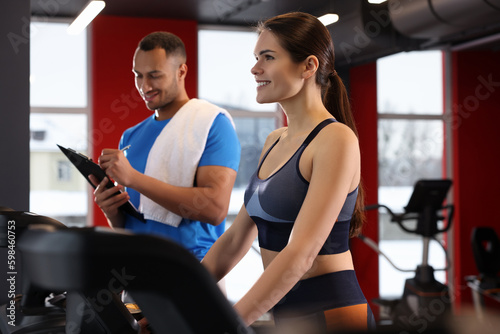 Happy trainer writing down workout plan while woman doing exercise in modern gym, space for text
