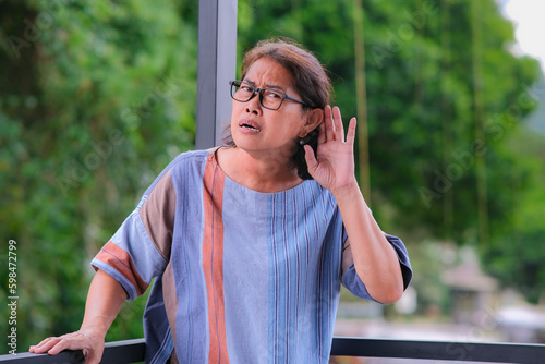 Asian middle-aged woman placing her hand on her ear to listen more clearly photo