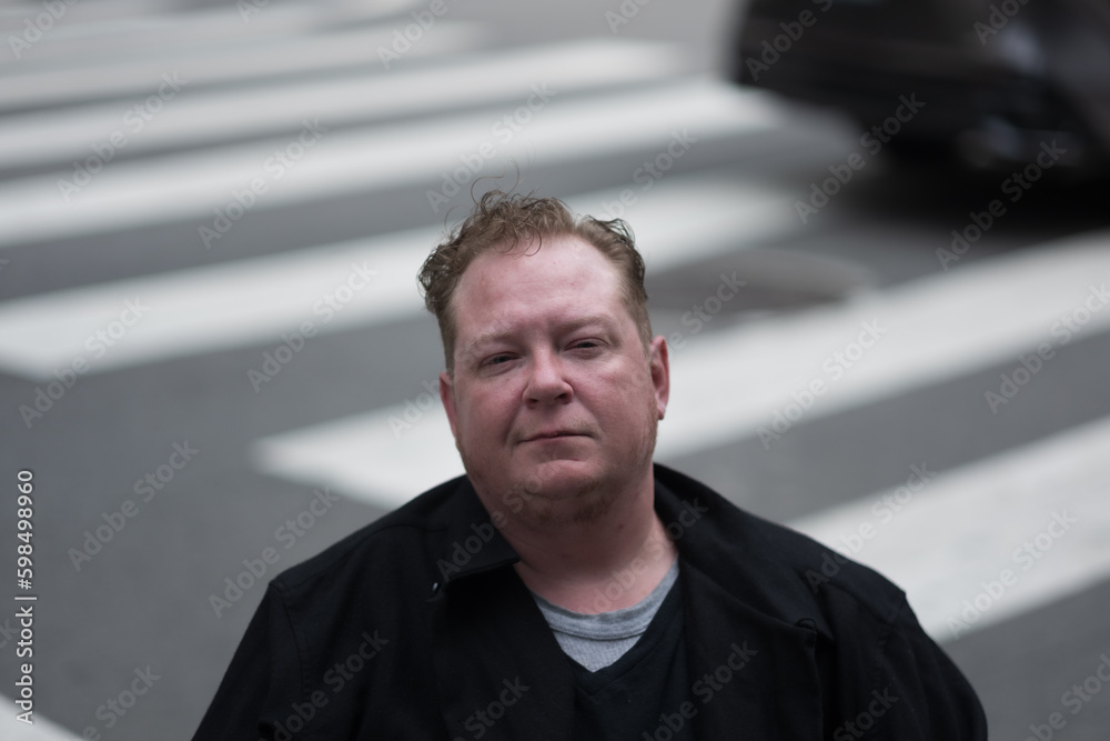 Looking directly at the camera, a disabled transman in a powerchair pauses in a crosswalk to express questions about movement and accessibility. There are zebra stripes and a car in the background.