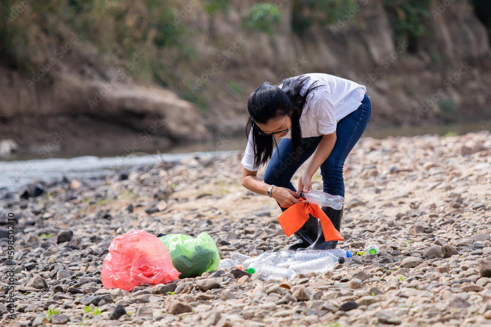 Young woman picking up trash by the stream, he concept of cleaning the coastal zone. Girl volunteer picks up a plastic bottle on the beach. Environment and pollution caring management concept.