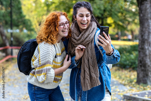 Two student woman friends in urban public park use mobile phone and smile