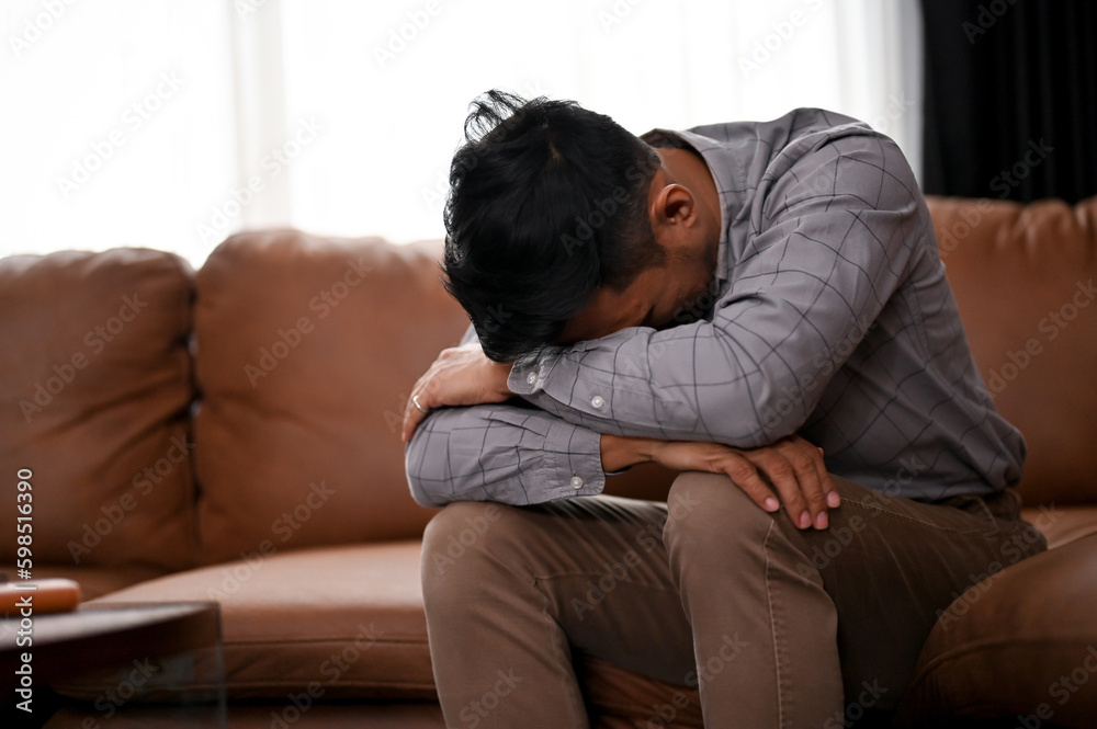 Stressed and depressed millennial Asian man is crying on the sofa in a living room