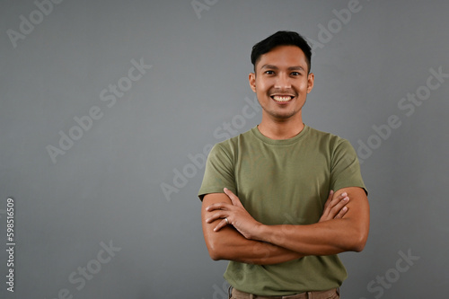 Handsome Asian man stands isolated on a grey studio background with his arms crossed.