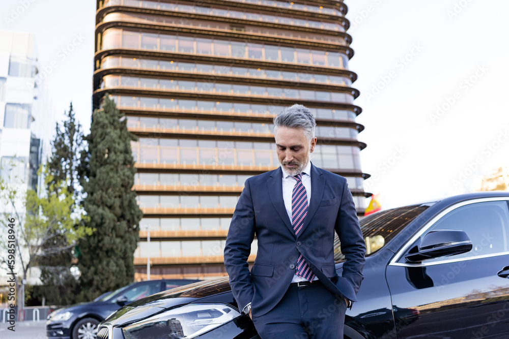 Businessman with hands in pockets looking down by car in front of building Businessman with hands in pockets looking down by car in front of building