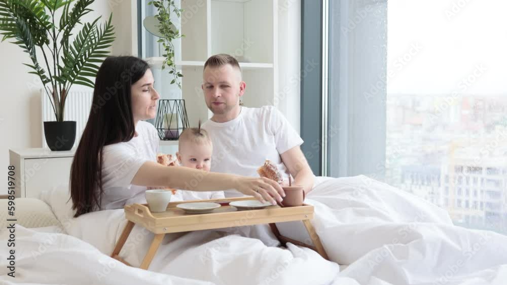 Sweet little baby drinking from sippy cup while joyful brunette woman snuggling with blonde male in cozy bedroom with tray on foreground. Caring family spending quality time together at home. Sweet little baby drinking from sippy cup while joyful brunette woman snuggling with blonde male in cozy bedroom with tray on foreground. Caring family spending quality time together at home.