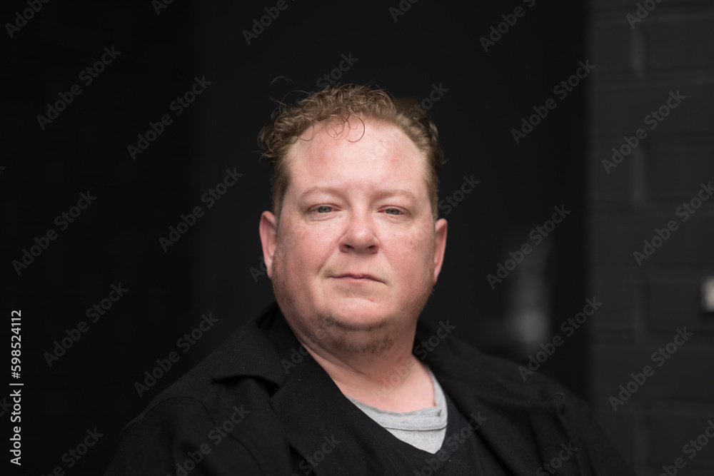 A disabled man celebrates his identity as a disabled and trans man, sitting in his powerchair with a black brick wall in the background, and expressing a direct, professional gaze. A disabled man celebrates his identity as a disabled and trans man, sitting in his powerchair with a black brick wall in the background, and expressing a direct, professional gaze.