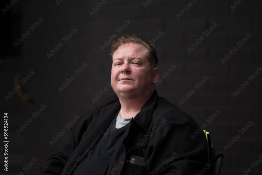 A proud disabled and trans man celebrates his identity in a powerchair against a black brick wall. He radiates optimism and confidence.
