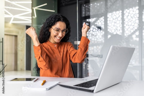 Hispanic business woman celebrating victory success, employee with curly hair inside office reading good news, using laptop at work inside office holding hand up and happy triumph gesture
