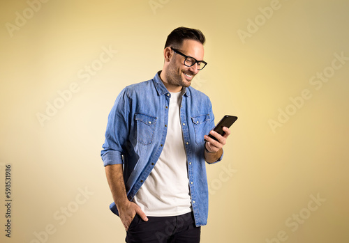 Charming male entrepreneur with hand in pocket smiling and messaging over mobile phone. Happy young man wearing denim shirt and eyeglasses standing isolated on yellow background © Moon Safari