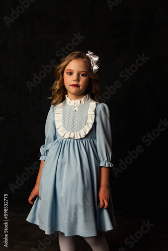 Sad little cover girl in image of doll in blue dress at black background, pensive looking at camera. Studio shot of cute child isolated with light and shadow. Kids emotion concept. Copy ad text space