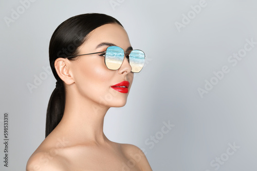 Attractive woman in stylish sunglasses on light grey background. Sandy beach and sea reflecting in lenses. Space for text