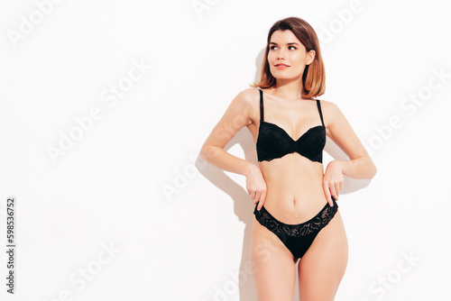 Young beautiful sexy woman in black lace lingerie. Carefree confident model wearing black underwear. Hot brunette posing on white wall in studio. Perfect body. Slim and fit. Isolated