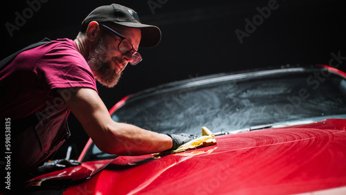 Car Wash Expert Using a Microfiber Cloth to Dry Up a Red Modern Sportscar. Adult Man Cleaning Away Dirt, Preparing an American Muscle Car for Detailing. Creative Cinematic Photo with Sport Vehicle photo