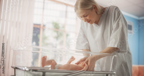 Mother Bonding with her Newborn Child Near Hospital Bassinet Bed in a Modern Nursery Clinic. Caring Mom Holding and Comforting Her New Born Child. Healthcare, Pregnancy and Motherhood Concept