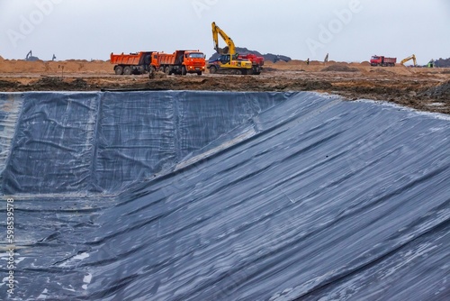 Ust-Luga, Leningrad oblast, Russia - November 16, 2021: Construction of chemical waste ditch (on front). Plastic cover of pit. Excavator and dump truck on the background