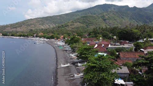Aerial drone view above seaside of Amed Village, Bali Indonesia. White jukung fishing boats on volcanic black sand beach,Villager fisher Houses, Traffic and tropical green mountains in background photo