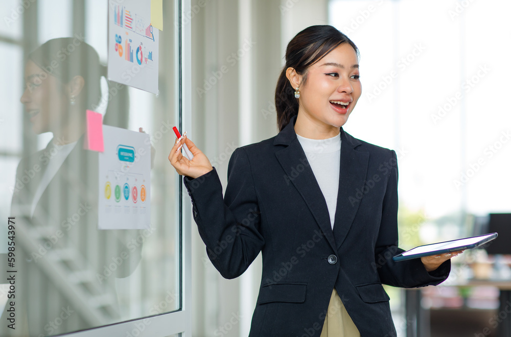 Asian professional successful female businesswoman lecturer presenter in formal business suit standing using marker pointing infographic paperwork document sticky note on glass board in meeting room Asian professional successful female businesswoman lecturer presenter in formal business suit standing using marker pointing infographic paperwork document sticky note on glass board in meeting room