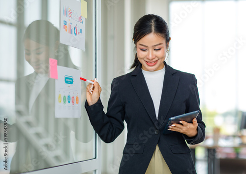 Asian professional successful female businesswoman lecturer presenter in formal business suit standing using marker pointing infographic paperwork document sticky note on glass board in meeting room