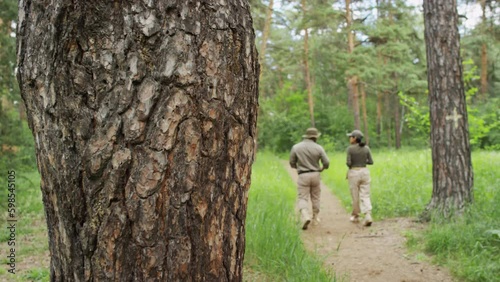 Full rear shot of man and woman in khaki forest ranger uniforms walking down pedestrian path in pine forest, engaged in discussion, and tree trunk with rugged bark in foreground photo