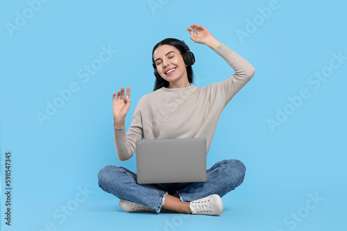 Happy woman with laptop listening to music in headphones on light blue background © New Africa