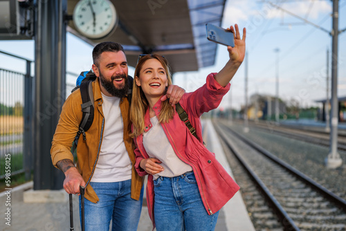 Happy couple is standing at railway station and waiting for arrival of their train. They are taking selfie.