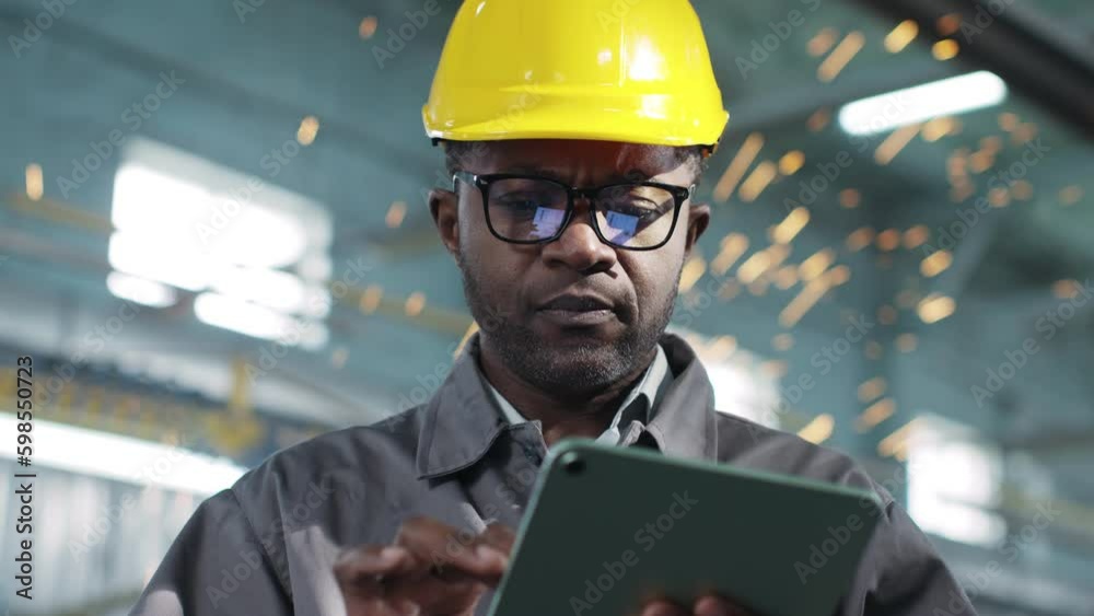 Man in glasses is calculating something on his gadget and is seriously thinking about something. African american manager in special uniform thinks about organization of work in workshop.