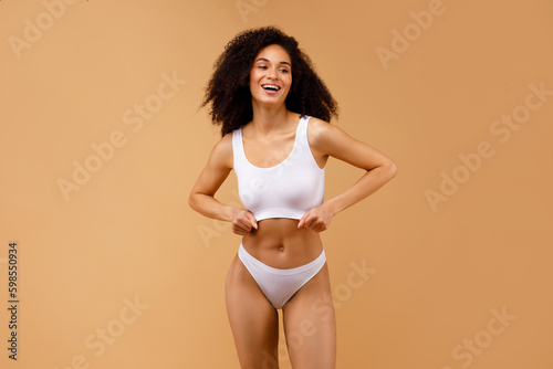 Slim african american woman in white underwear top bra and panties standing isolated over beige studio background Generative AI