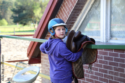 The girl is riding a horse. He puts on a helmet, unsaddles and saddles a horse, hangs a saddle, rides a white horse in an arena. Active recreation and sports, happy child photo