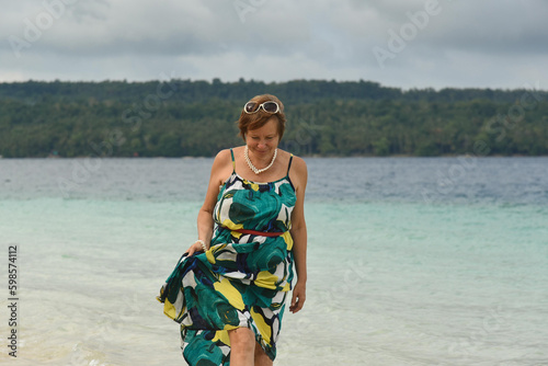 Happy attractive woman walks and dances on the beach. Senior woman wear the green long dress and sunglasses. Concept of active retired elderly and travel lifestyle. Landscape on the background.