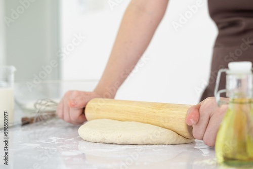 Pizza dough with ingredients on white table. Woman hands keep rolling pin with flour in white bright kitchen, baking background, menu, recipe. Preparing bread dough in kitchen at home.