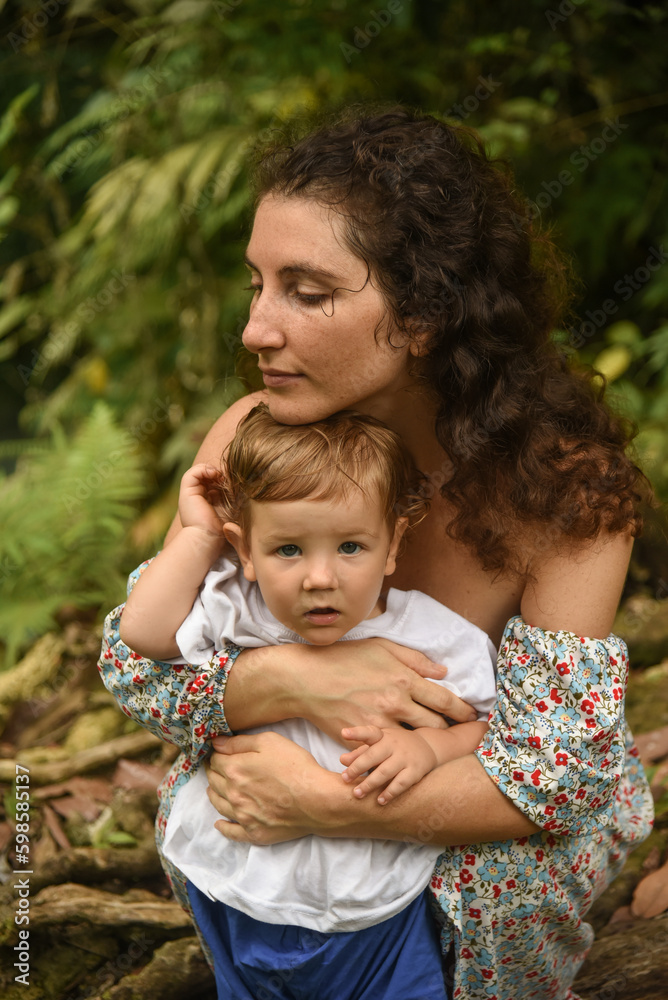 Thoughtful curly mother in a dress with a baby spend time in nature. Cute family in the summer city park. Mother hugs her son. Thoughtful curly mother in a dress with a baby spend time in nature. Cute family in the summer city park. Mother hugs her son.