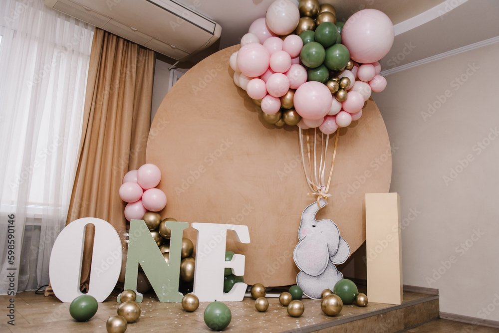 Arch decorated pink, golden, green balloons, paper decor elephant, confetti. Birthday party for 1 year old girl or boy on background brown photo wall. Reception. Celebration concept. Space for text.