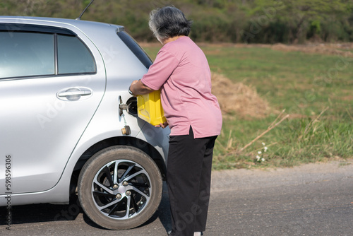 The car ran out of gas and stalled beside the road in suburbs and an elderly Asian woman used a gallon of spare gas to fuel the car. A woman prepares a gallon of spare gas to fuel before traveling.